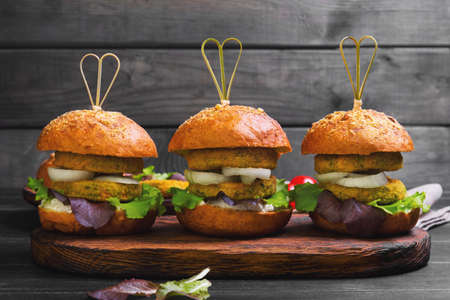 Vegan burgers with vegetables, onion, vegetable cutlets, lettuce, sauce, ingredients for cooking vegan burgers cherry tomatoes, lettuce, rolls, skewers on wooden surface of dark black backgroundの写真素材