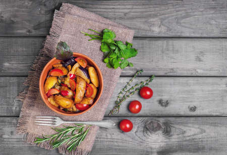 Steamed vegetables with rustic roasted potatoes, eggplant, zucchini, peppers, fresh cherry tomatoes, rosemary, cilantro, basil, thyme in ceramic bowl on light background gray wooden worn, top viewの写真素材