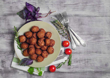 Small meat balls on green plate, fresh special spices for meat balls herbs thyme, coriander, basil, rosemary, cherry tomatoes, silver fork on light white wooden background, rustic style, top viewの写真素材