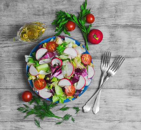 Healthy vegetarian food, fresh vegetables salad with different vegetables radishes, cherry tomatoes, lettuce salad, dill, sauce olive oil, fresh whole radishes,  blue plate, light wooden background, top viewの写真素材