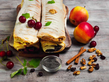 Assortment poppy seed strudel, apple and cinnamon strudel, cherry strudel, cheese raisin strudel, fresh cherries, apples, poppy seeds, raisins, walnuts, lemon balm, tray, light white wooden backgroundの写真素材