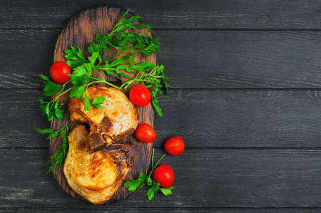 Grilled meat pork chops, served in grill board, ingredients for meat pork chops, dill, parsley, lettuce, cherry tomatoes, dark black wood background, top viewの写真素材