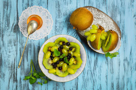 Fruit and berry salad. Ingredients for salad of kiwi, blueberry, grape, Melissa, honey. Kiwifruit on white saucer. Blue wood background. Top view.の写真素材