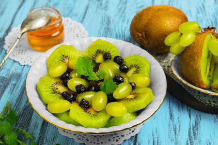 Fruit and berry salad. Ingredients for salad of kiwi, blueberry, grape, Melissa, honey. Kiwifruit on white saucer. Blue wood background.の写真素材