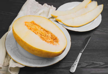 Fresh ripe yellow melon on white porcelain plate, three pieces of sliced melon, knife, cloth, dark brown wooden backgroundの写真素材