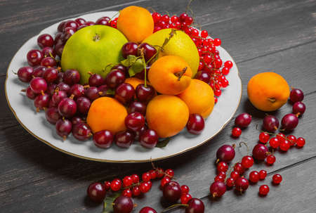 White plate with an assortment of fresh garden fruits and berries. Fruits apricot, apple, pear. Berries red currants, cherries, gooseberries. On dark black wooden table fruit and gooseberries, apricots, red currants, cherries.の写真素材