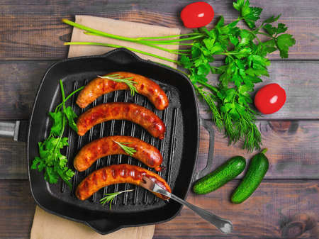 Grilled fried sausages kupaty on a cast iron skillet grill, greens to grilled fried sausages parsley, thyme, rosemary, tomatoes, cucumbers. Fork for fried sausages. Grey wooden background. Top view.の写真素材