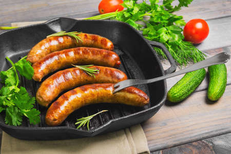 Grilled fried sausages kupaty on a cast iron skillet grill, greens to grilled fried sausages parsley, thyme, rosemary, vegetables, tomatoes, cucumbers. Fork for fried sausages. Grey wooden background.の写真素材