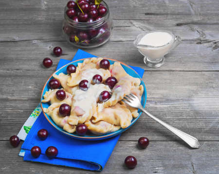 In blue ceramic dish, fruit and berry dumplings with cherries. Fresh cherry dumplings for bank. Sour cream for dumplings with cherries. Gray wooden background.の写真素材