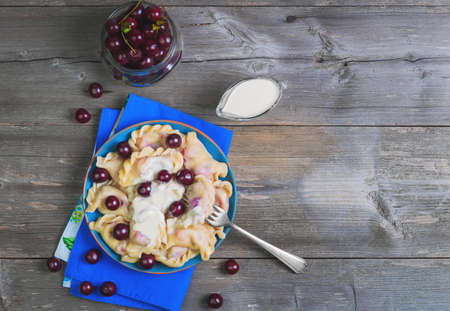 In blue ceramic dish, fruit and berry dumplings with cherries. Fresh cherry dumplings for bank. Sour cream for dumplings with cherries. Gray wooden background. Top viewの写真素材