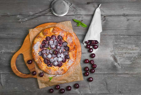 Open pie tart dough sprinkled powdered sugar with ripe cherries on paper on board, fresh cherry in white paper bags, red cherry for pie tart on gray wooden background. Top view.の写真素材