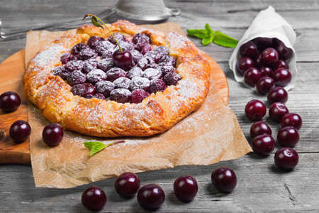 Open pie tart dough sprinkled powdered sugar with ripe cherries on paper on board, fresh cherry in white paper bags, red cherry for pie tart on gray wooden background.の写真素材