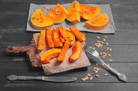Halloween Baked pumpkin pieces on wooden board, raw pumpkin pieces cut for baked, napkin tissue, pumpkin seeds on dark black backgroundの写真素材