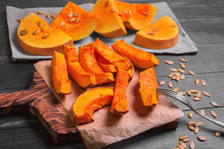 Halloween Baked pumpkin pieces on wooden board, raw pumpkin pieces cut for baked, napkin tissue, pumpkin seeds on dark black backgroundの写真素材