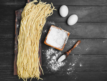 Homemade Uncooked pasta spaghetti noodles on board. Ingredients for home-made spaghetti noodles chicken eggs, flour. The dark black wood background. Top view, blank space.の写真素材