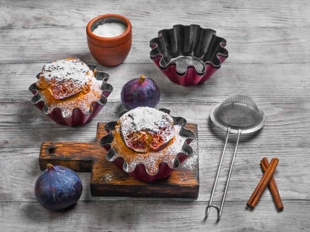 Baked Muffins cake with figs, sugar powder. Fresh fruit muffin figs. Metal molds for muffins, cinnamon, board. Light white wooden background.の写真素材