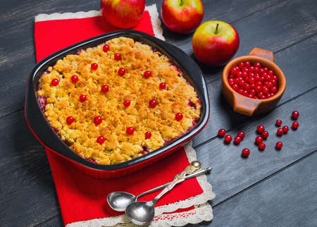 Dessert pie crumble cake apples, red berries cranberries. Fresh apples, berries for  pie crumble cake, cloth. Red Black ceramic baking pan cake crumble. Wooden dark background.の写真素材