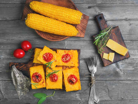 Pieces Corn polenta baked with cherry tomatoes, Parmesan cheese on board. Boiled corn on wooden plate for polenta. Green for corn polenta thyme, rosemary. Gray wooden background. Top viewの写真素材