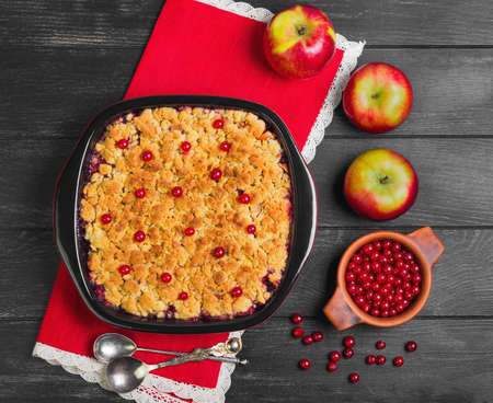 Dessert pie crumble cake apples, red berries cranberries. Fresh apples, berries for  pie crumble cake, cloth. Red Black ceramic baking pan cake crumble. Wooden dark background. Top view.の写真素材