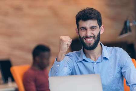 Young bearded caucasian modern business man sitting in a startup office using laptop, looking downward the screen, smiling - business, work, technology concept.の写真素材