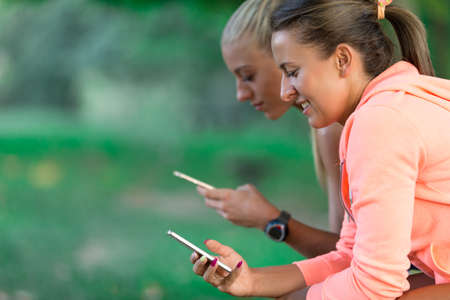 Pretty young woman in sporty outfit using a smartphone and listening to music while sitting on a park benchの写真素材
