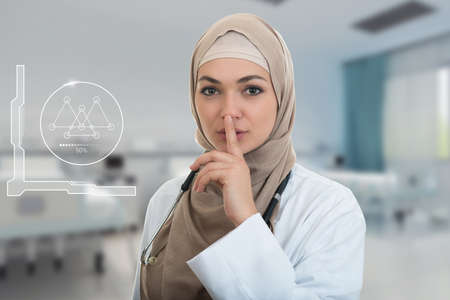 Closeup portrait of friendly, confident muslim with hijab doctor showing shh sigh, silence, healthcare professional in modern hospitalの写真素材