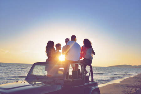 Five young people having fun in convertible car at the beach at sunset. They are wearing summer...