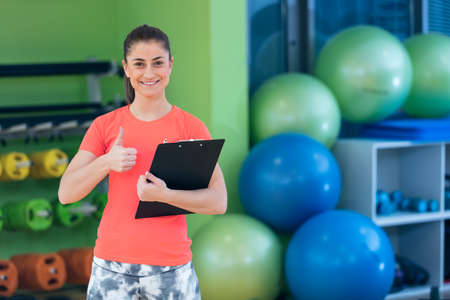 Portrait of smiling female fitness instructor writing in clipboard while standing in gym