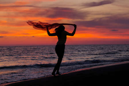 Silhouette of young girl, jumping with silk cloth against of sea sunsetの写真素材