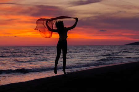 Silhouette of young girl, jumping with silk cloth against of sea sunsetの写真素材