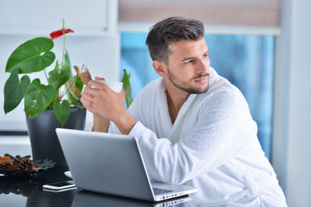 Handsome young man drinking coffee while working with laptop in kitchenの写真素材