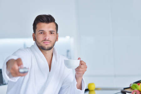 Young man in bathrobe sitting on kitchen worktop and having a cup of teaの写真素材