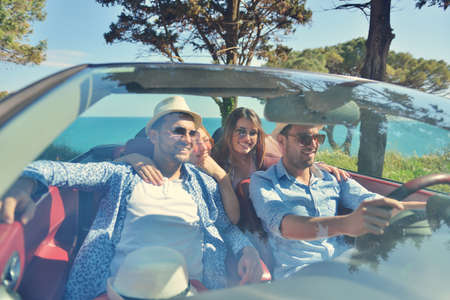 leisure, road trip, travel and people concept - happy friends driving in cabriolet car along country...