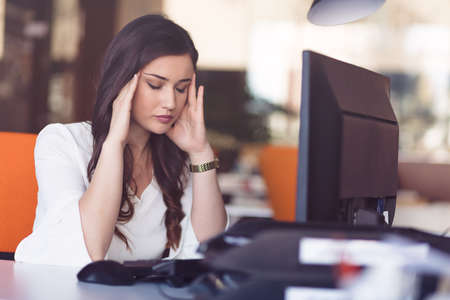 Young tired business woman with headache sitting in workplaceの写真素材