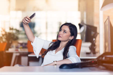 Beautiful young woman is making selfie in her workplace. She is relaxing and putting her legs on the table.の写真素材