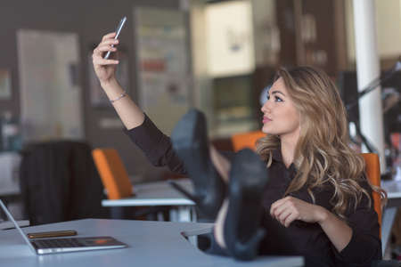 Beautiful young woman is making selfie in her workplace. She is relaxing and putting her legs on the table.の写真素材