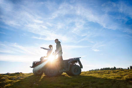 Beautiful couple is watching the sunset from the mountain sitting on quadbikeの写真素材