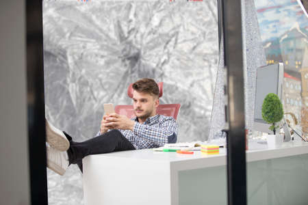Handsome businessman in classic shirt is using a smartphone and smiling while sitting with legs on table in officeの写真素材