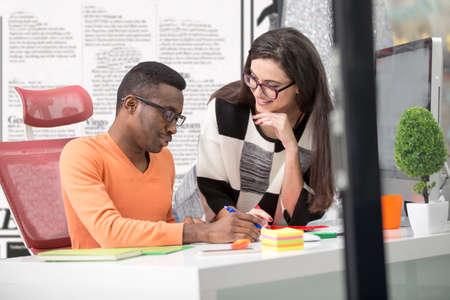 Two diverse work colleagues smiling and writing down notes while sitting together at a table in a...
