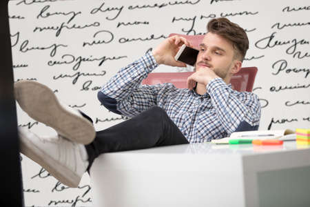 Handsome businessman in classic shirt is using a smartphone and smiling while sitting with legs on...
