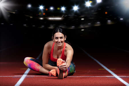 young athletic woman doing exercises on flexibility. stretching legs sitting track stadium, night with reflectorsの写真素材