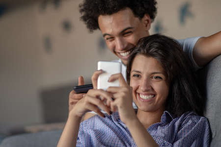 Attractive couple sitting on couch together looking at smartphone at home in the living roomの写真素材