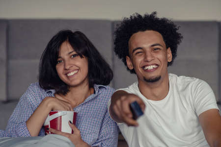 Image of smiling couple sitting on floor near sofa at home and looking aside while eating popcorn