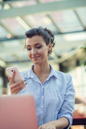 Portrait of a playful young girl taking selfie with mobile phone while sitting with laptop computer at a cafe outdoorsの写真素材