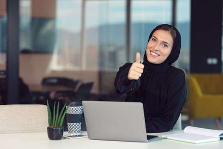 Young arabic businesswoman sitting at workplace in office showing thumbs upの写真素材