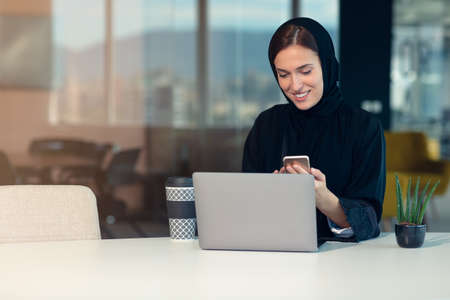 Happy muslim businesswoman in hijab at office workplace. Smiling Arabic woman working on laptop and talking on smartphoneの写真素材