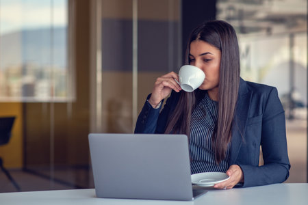 Young woman working on laptop and drinking coffee in officeの写真素材