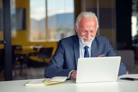 Handsome smiling senior man while sitting at his cozy workplace with laptop at home, retired male chatting with friends in social media,の写真素材