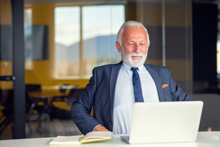 Handsome smiling senior man while sitting at his cozy workplace with laptop at home, retired male chatting with friends in social media,の写真素材