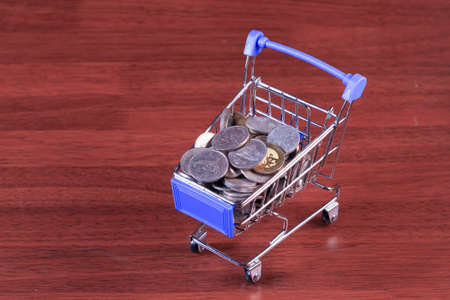 Miniature shopping cart full of coins on a wooden background- payments, investments and commerce conceptの写真素材
